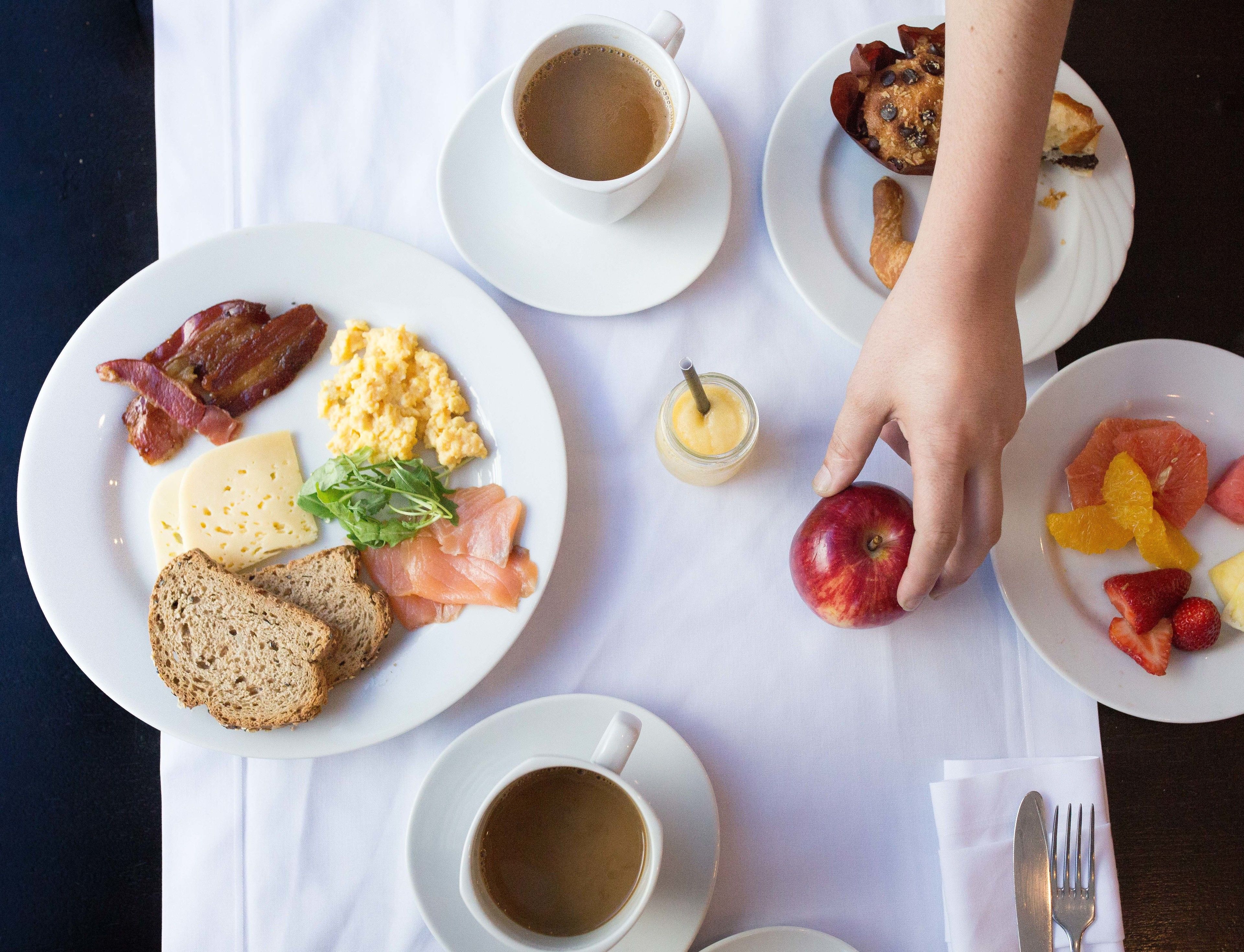 Una mesa de desayuno con platos de comida, tazas de café y una mano alcanzando una manzana.