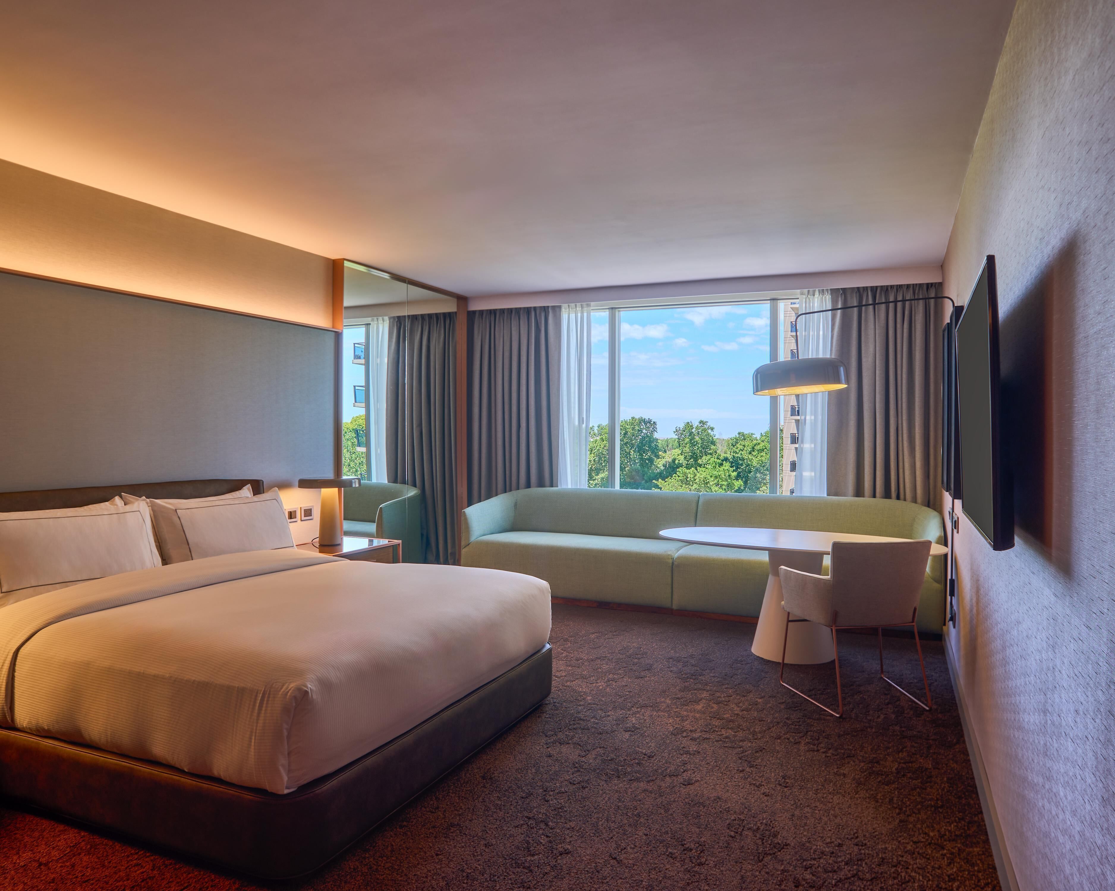 Quarto de hotel com cama, sofá, mesinha, televisão e janela com cortinas.