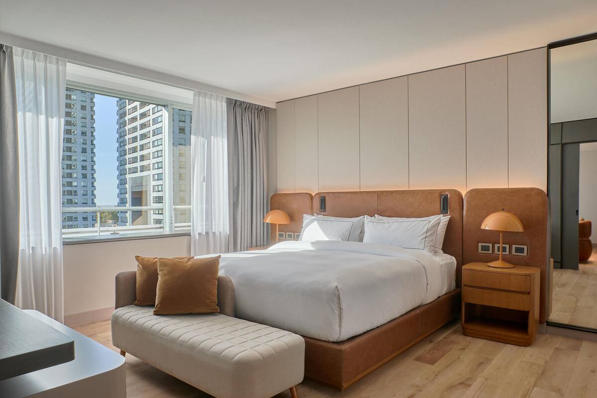 Ett modernt hotellrum med en stor säng, ett sängbord, en liten soffa och utsikt över höga byggnader genom fönstret.