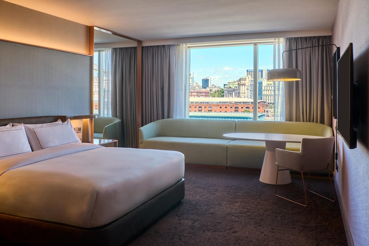 Ett hotellrum med en säng, en soffa, ett litet bord, en stol, en tv och ett stort fönster med utsikt över staden.
