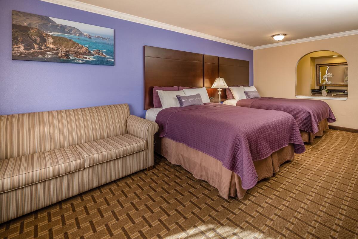 Une chambre d’hôtel avec un canapé à rayures, deux lits avec une literie violette, un tableau représentant un paysage côtier et une moquette à motifs.