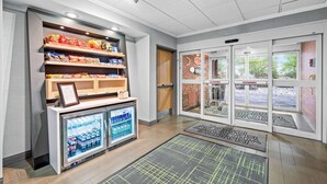 A clean, modern lobby with a refrigerated display case, shelves with snacks, and a glass door entrance. - Hampton Inn Lincoln - South/Heritage Park (Lincoln)