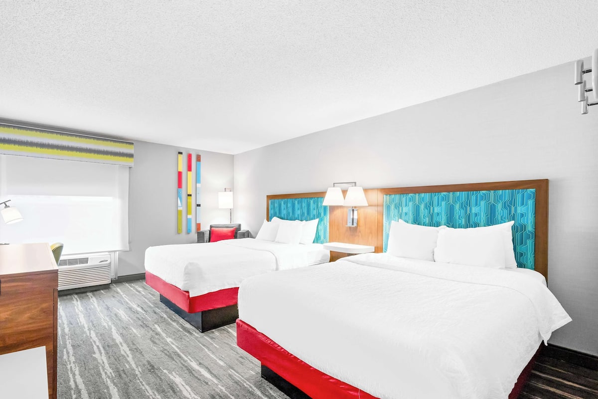 A hotel room with two beds, a desk, a chair, a window with blinds, and a wall-mounted air conditioner.