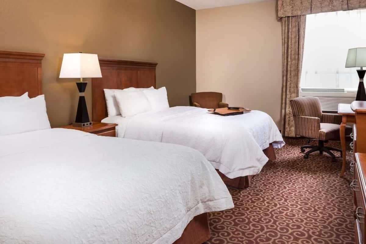 A hotel room with two beds, a desk, a chair, a lamp, and a window with curtains.