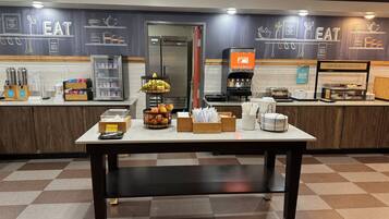 A breakfast area with a coffee machine, fruit display, and various breakfast items on a counter.