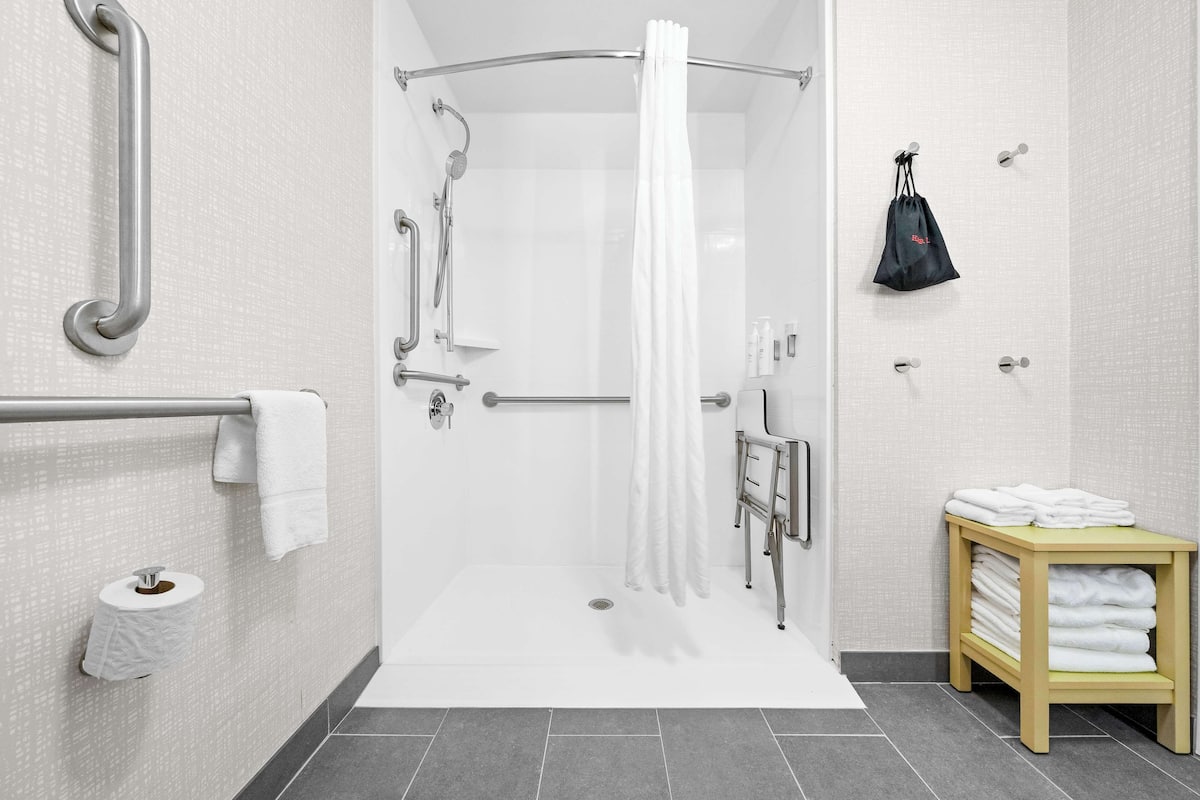 A modern bathroom with a shower, grab bars, a toilet paper holder, a towel rack, and a small table with towels.