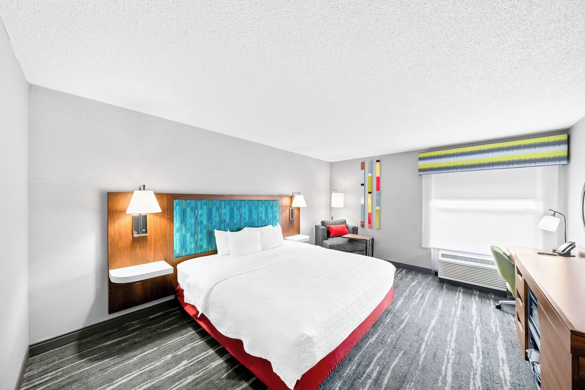A hotel room with a large bed, bedside tables, a desk, and a window with blinds.