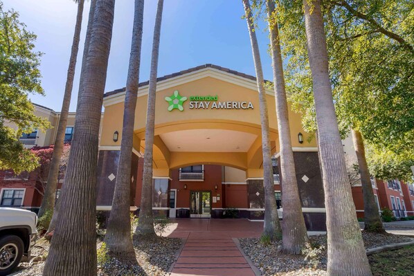 Exterior de um hotel Extended Stay America, com entrada central, palmeiras e céu claro.