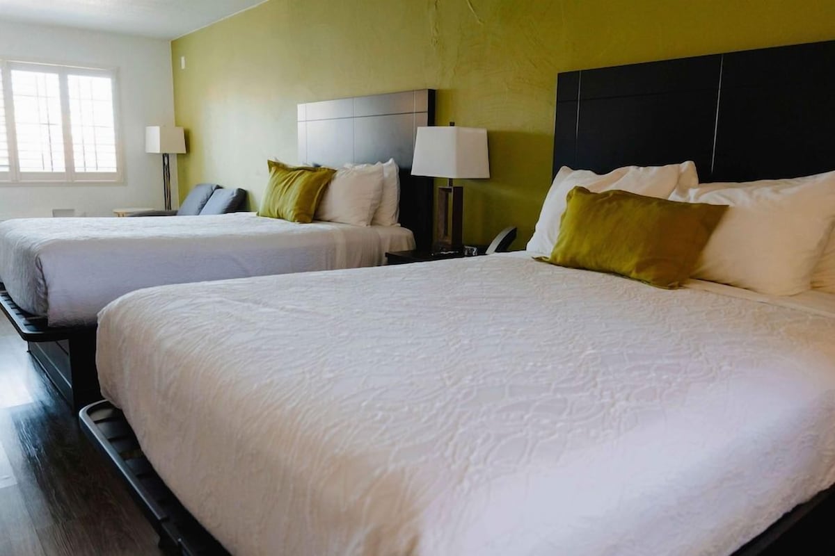 A hotel room with two beds, each with white linens and pillows, a nightstand with a lamp, and a window providing natural light.