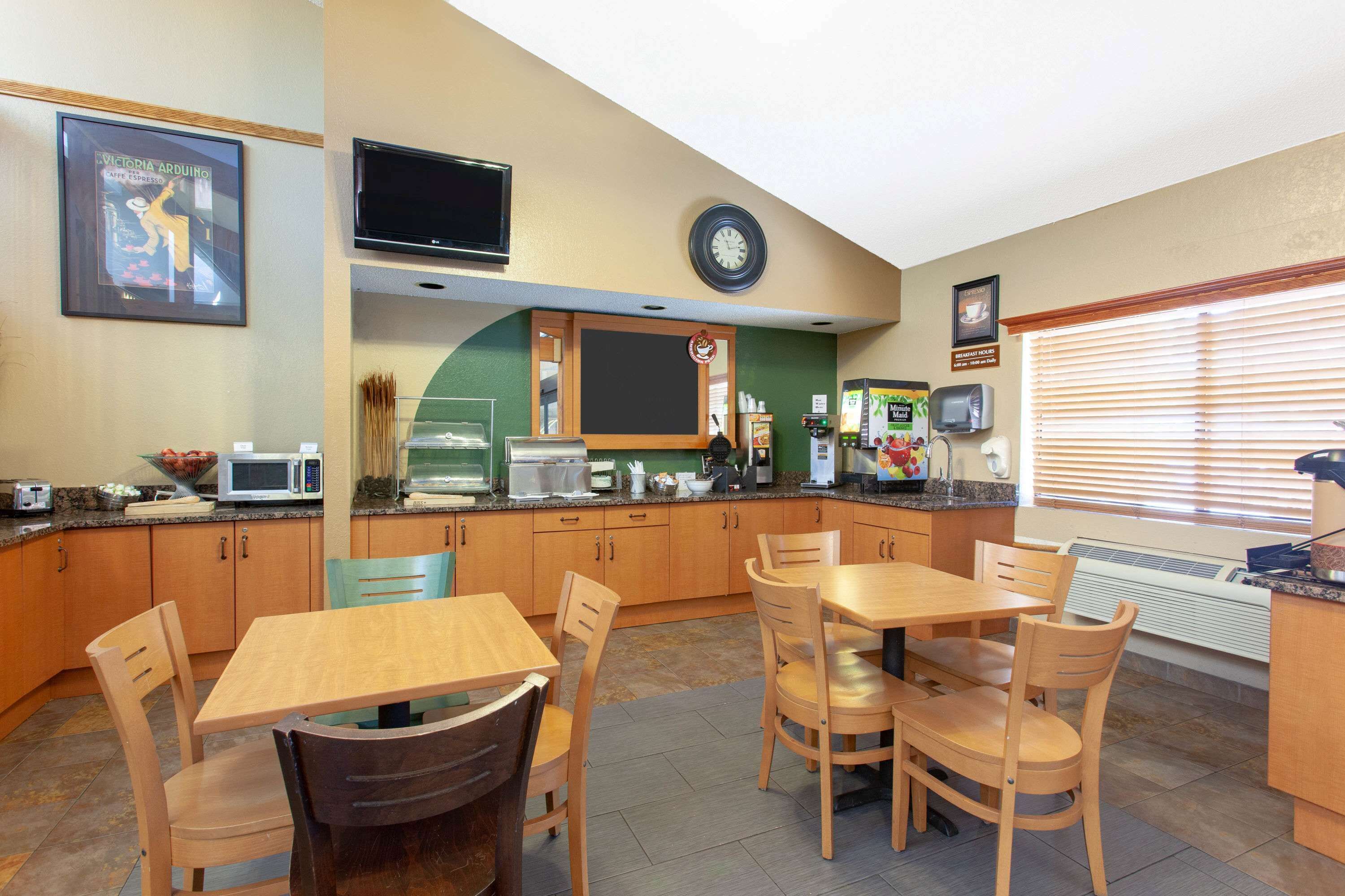 A modern kitchen with wooden cabinets, a dining table, and chairs. There is a microwave, toaster, and coffee maker on the counter. A television is mounted on the wall above the counter.
