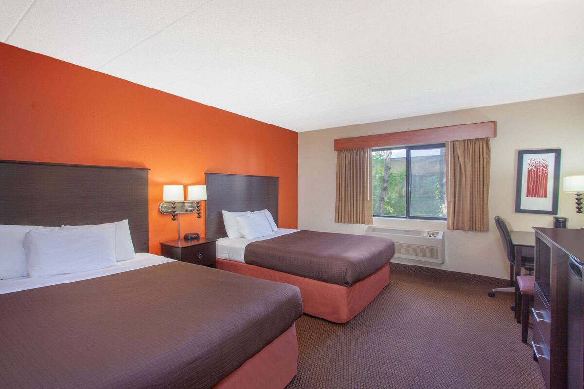 Habitación de hotel con dos camas, un escritorio, una silla, una ventana con cortinas y un cuadro en la pared.