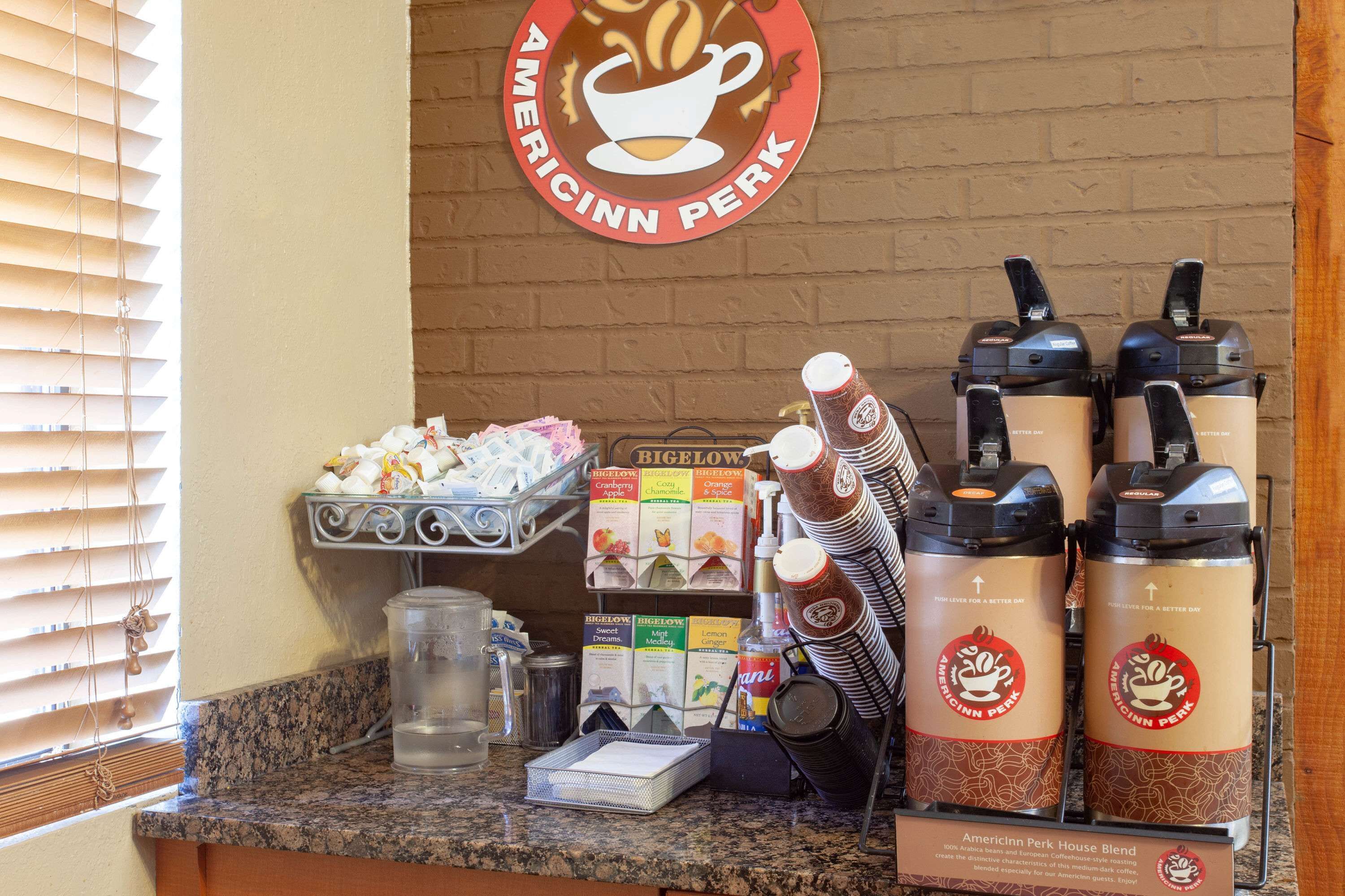 A coffee station with Americinn Perk branded coffee makers, a Bigelow coffee grinder, and various coffee pods and milk cartons.