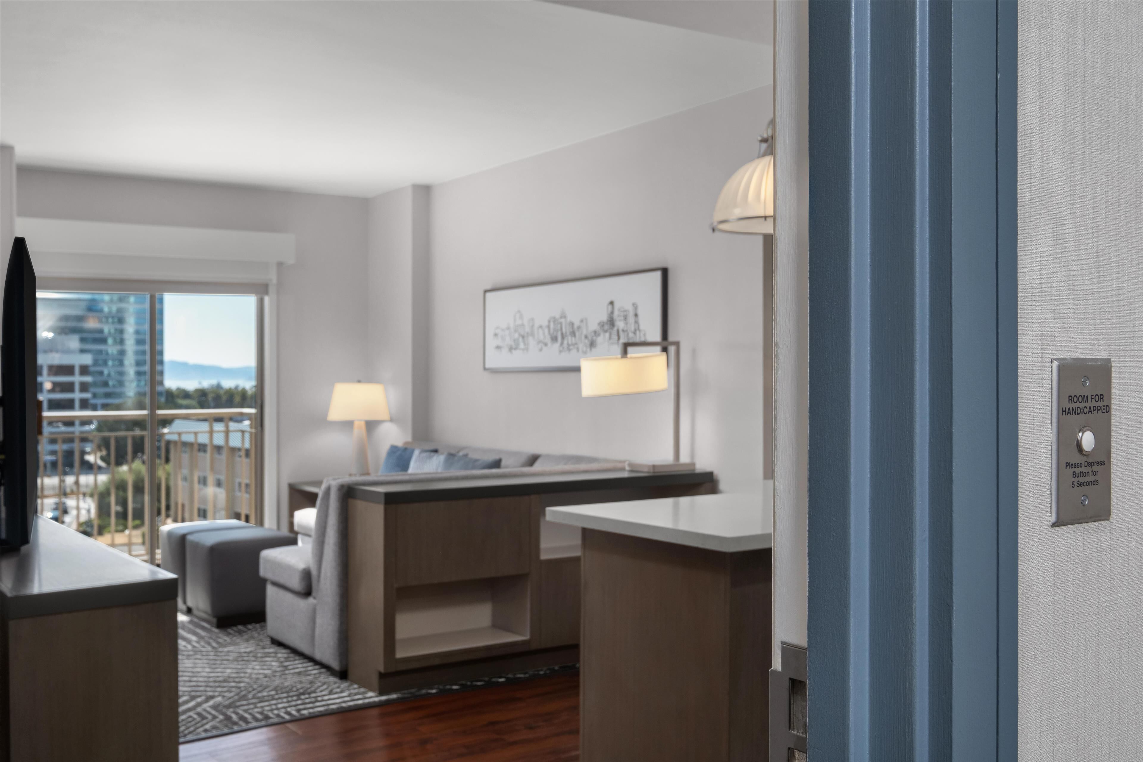 Una camera d'albergo moderna con divano, un tavolino, una lampada e vista sulla città attraverso ampie finestre.