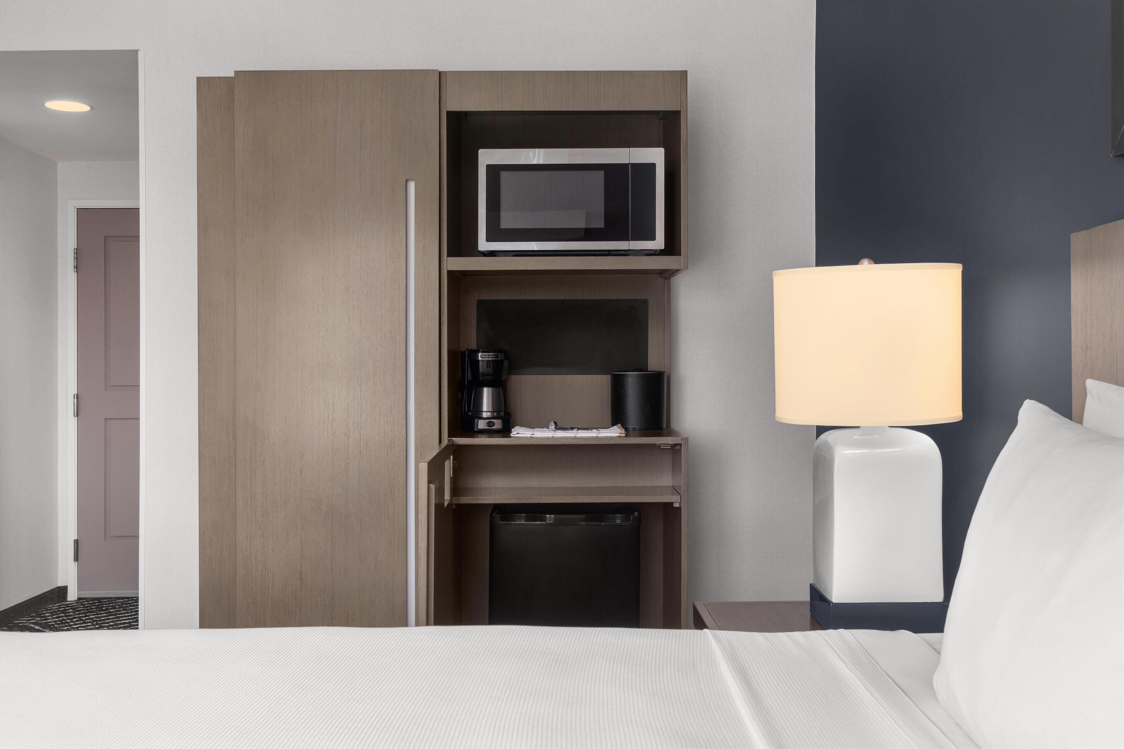 A hotel room with a bed, a bedside lamp, a microwave, a coffee maker, and a small cabinet.