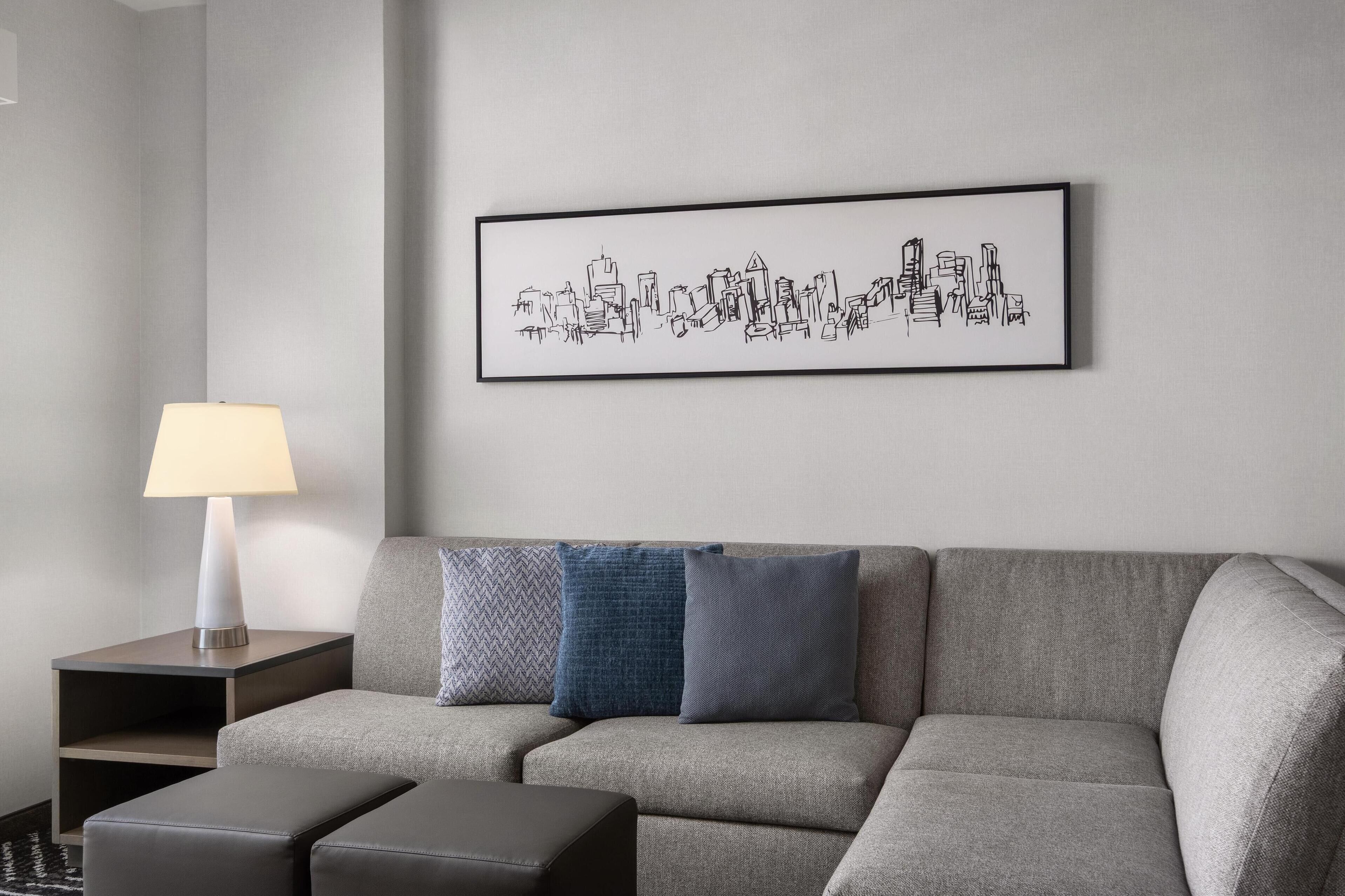 Una sala de estar moderna con un sofá gris, una mesita de noche con lámpara y una obra de arte con un paisaje urbano enmarcado en la pared.