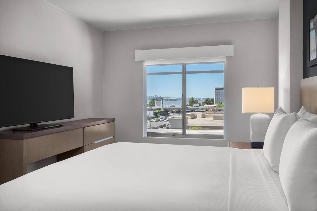 A modern hotel room with a large bed, a flat-screen TV, a desk, and a window offering a city view.