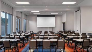 A conference room with multiple tables and chairs, a large screen, and windows providing natural light.