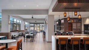 Un comedor moderno con varias mesas y sillas, una zona de cocina con asientos tipo barra, y ventanas grandes que dejan entrar luz natural.