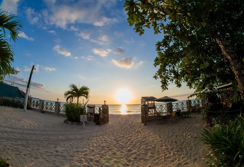 Une plage avec une clôture en bois, un espace de repos abrité et une vue sur le coucher de soleil sur l’océan.