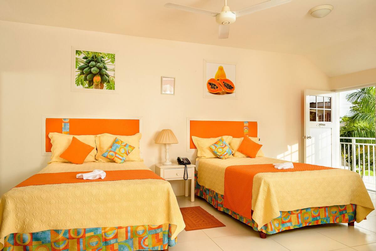A hotel room with two beds, each with orange bedding and colorful pillows, a small bedside table with a lamp, and a ceiling fan.