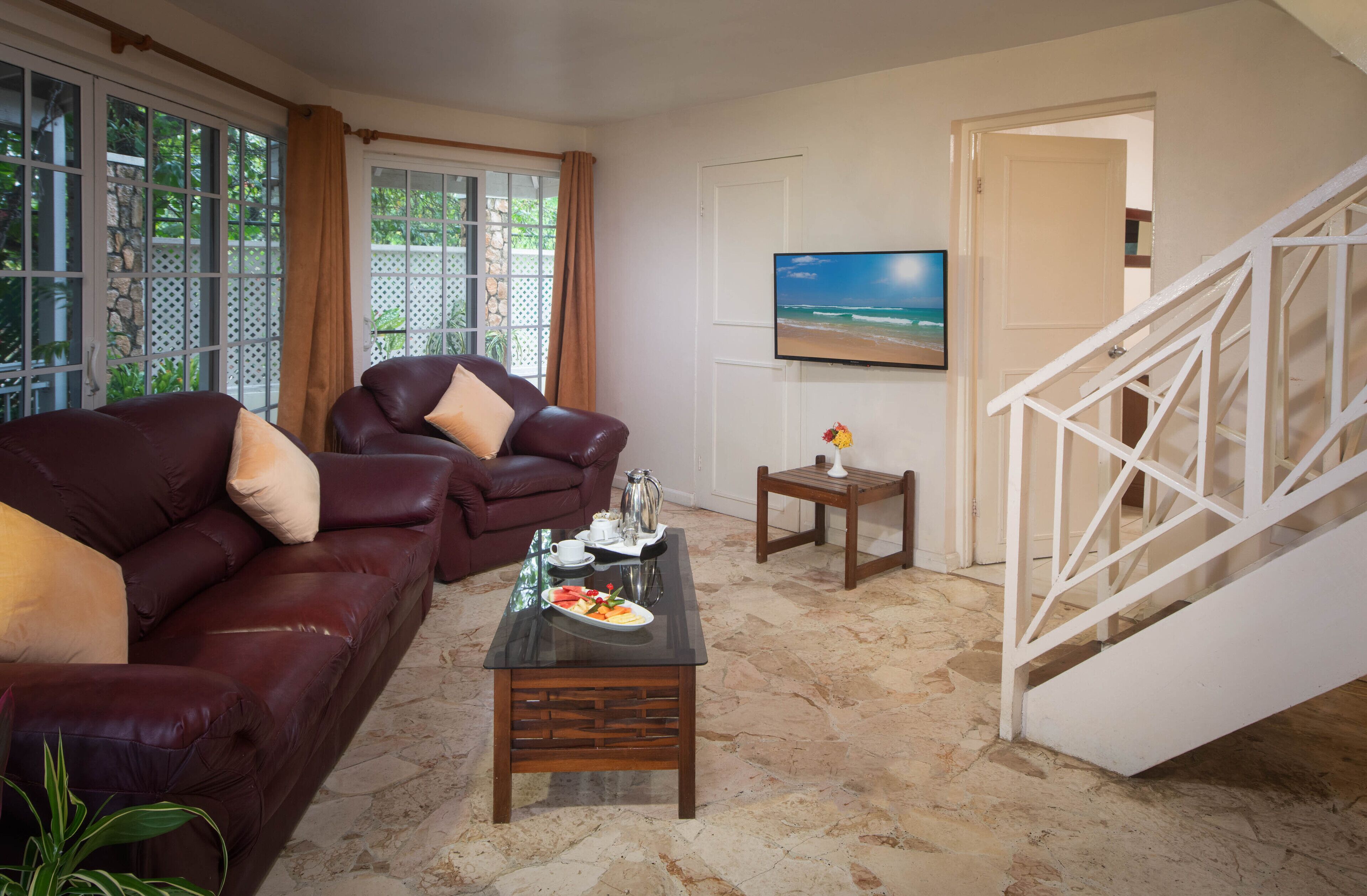 A living room with a leather sofa, a coffee table with a plate of food, a TV mounted on the wall, and a staircase.