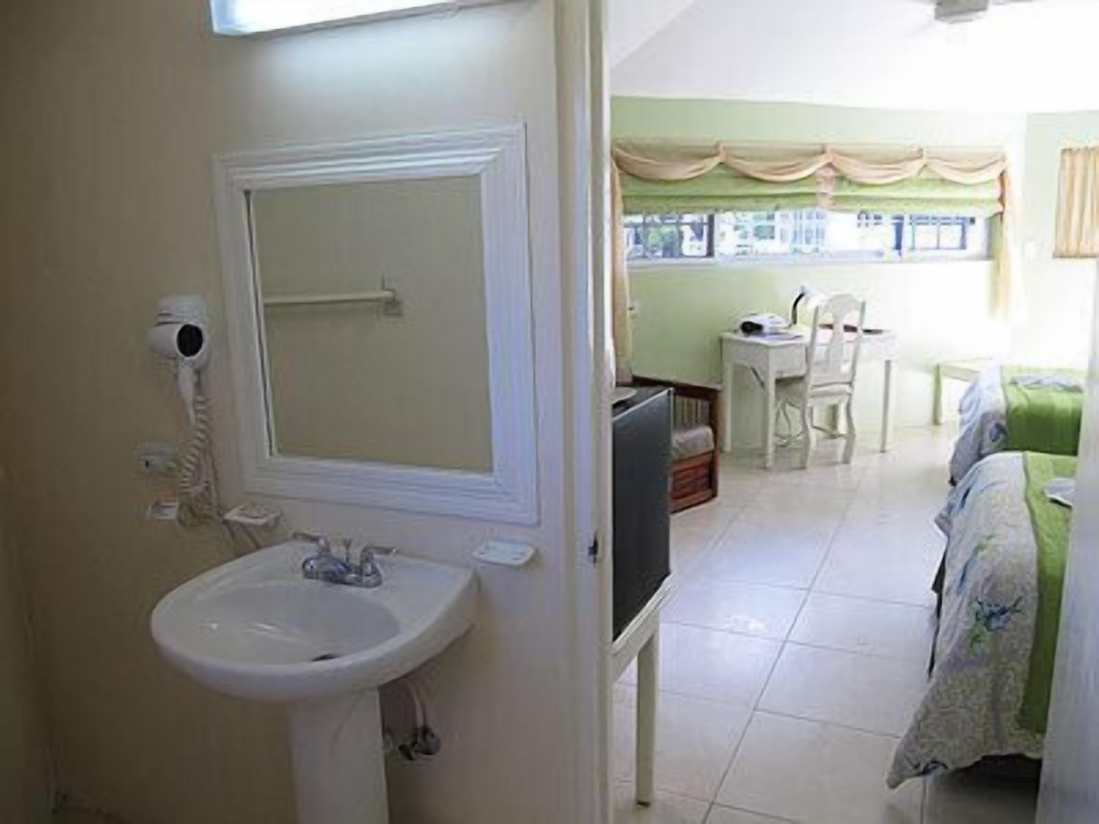 A bathroom with a sink, mirror, and hairdryer. There is a bed with a green blanket and a chair in the adjacent room.