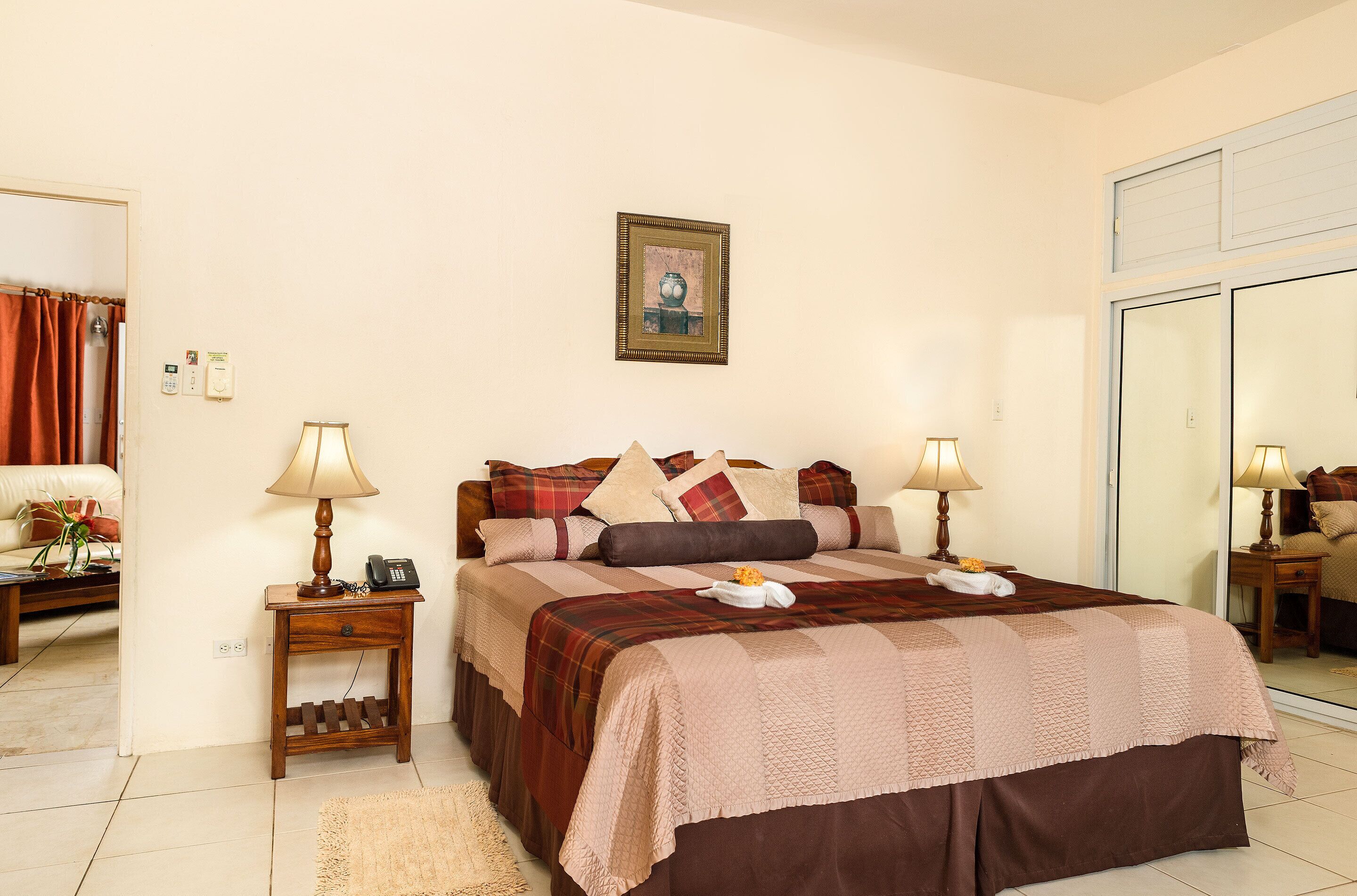 A neatly arranged bedroom with a large bed, two bedside tables with lamps, a bedside phone, a small shelf, and a framed picture on the wall.