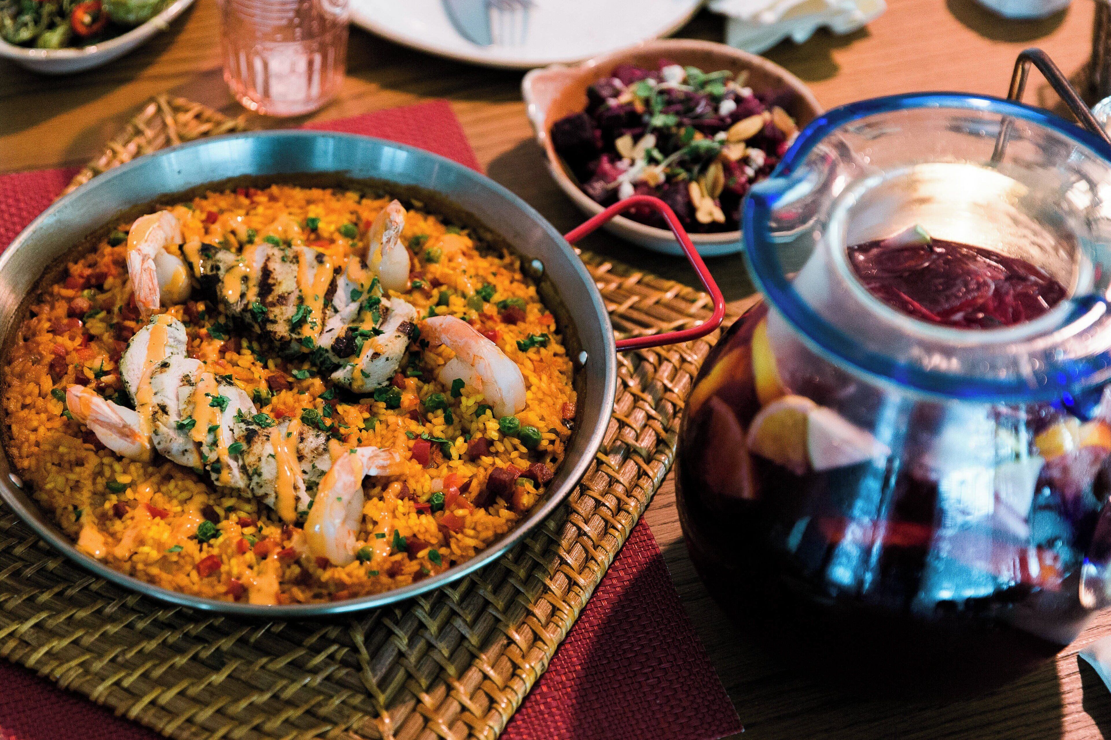 A large pan of shrimp and rice dish on a woven placemat, a bowl of salad, and a glass pitcher with a red handle.