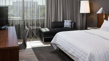 Een moderne hotelkamer met een bed, bureau, stoel, bank en stadszicht door grote ramen.