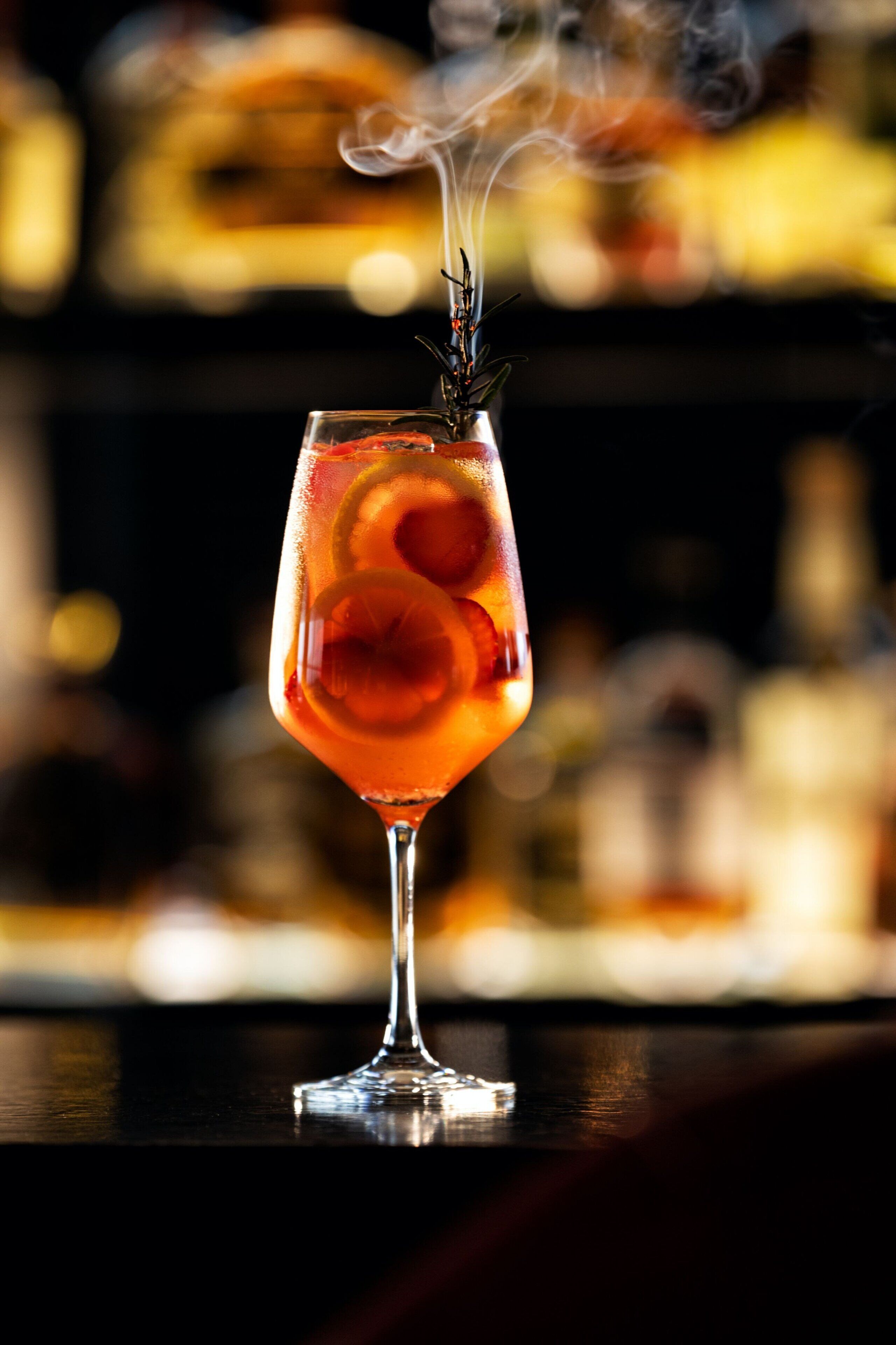 Een cocktailglas met garnering, damp die opstijgt, en een onscherpe achtergrond van flessen.