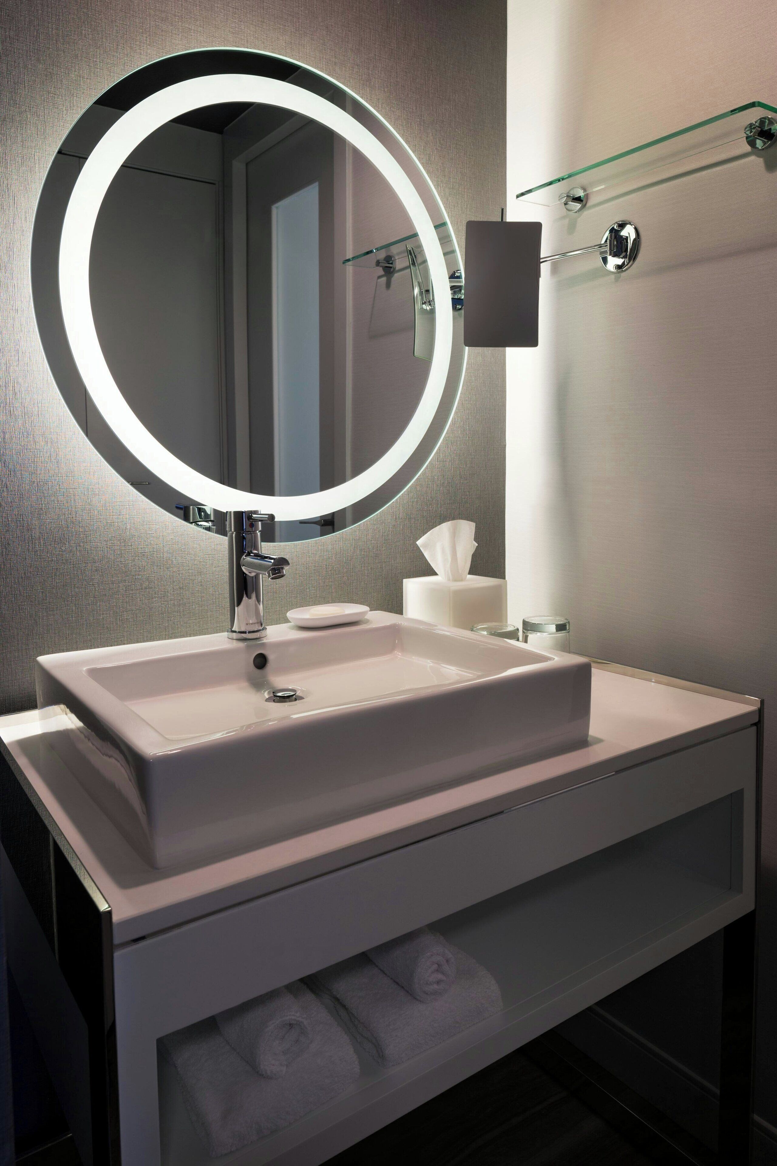 A modern bathroom with a large mirror, a white sink, and a glass shelf.