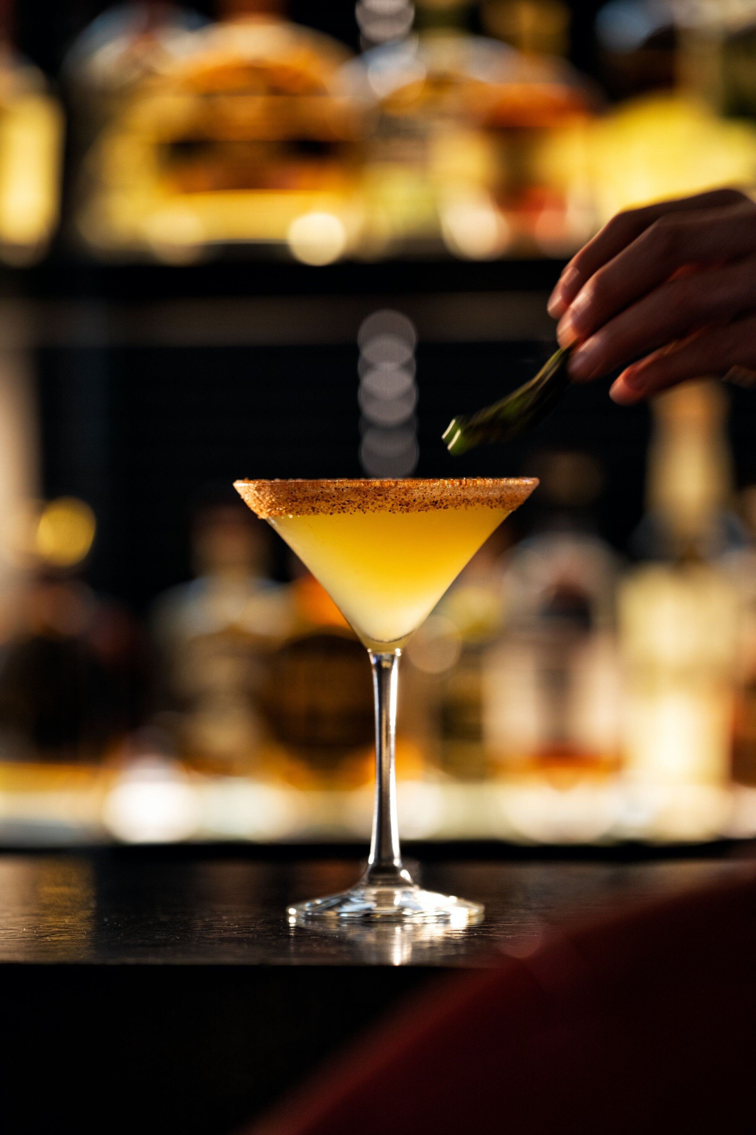 A cocktail in a martini glass with a salted rim, a hand holding a green garnish, and a blurred background of bottles.