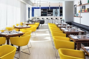 A restaurant with yellow chairs, wooden tables, and a bar area in the background. - Le Méridien Chicago - Oakbrook Center (Oak Brook)