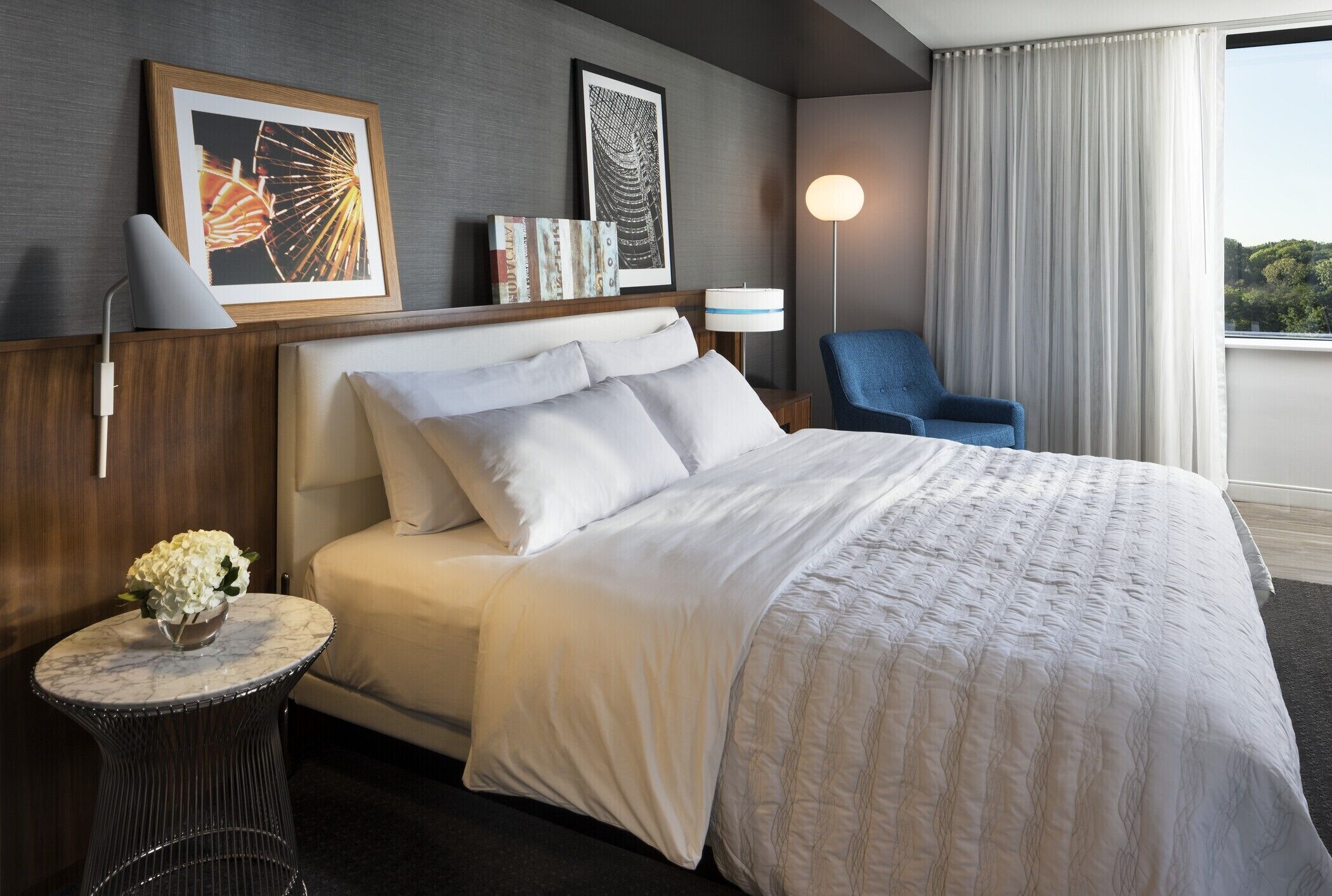 A neatly made bed with white linens, a small round table with a vase of flowers, and a blue chair.