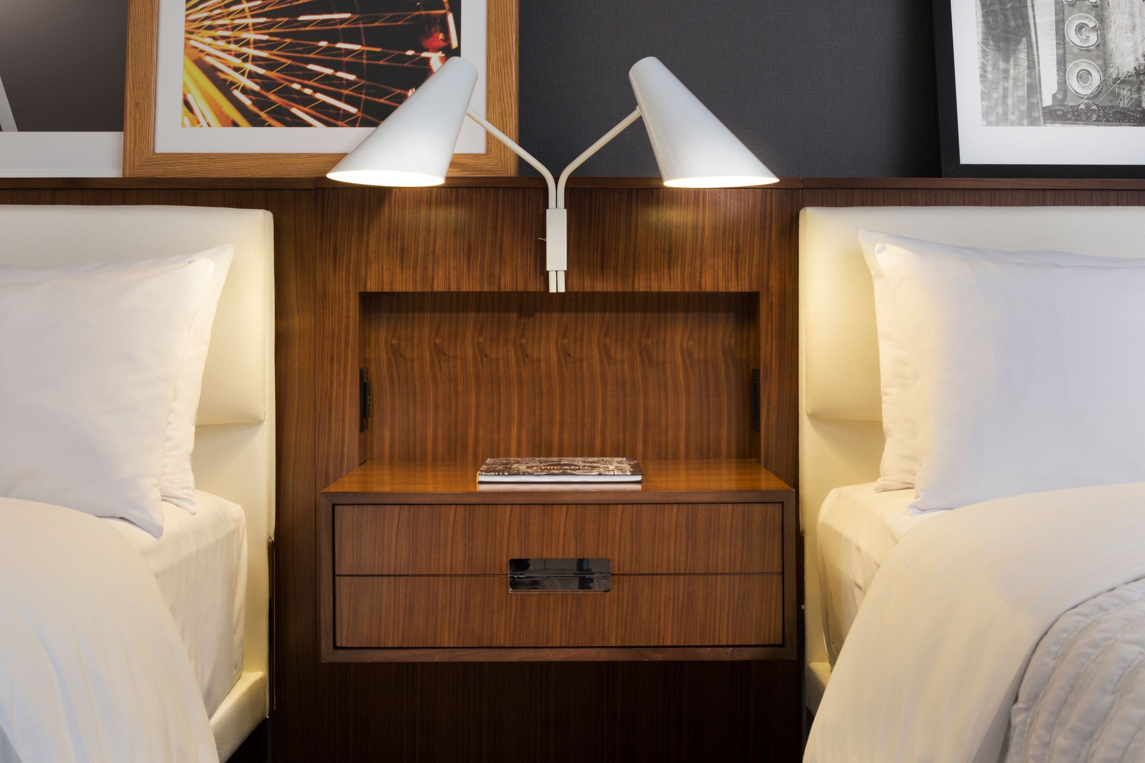 Una cama bien hecha con ropa de cama blanca, una mesita de noche de madera con un cajón y una lámpara blanca con dos pantallas.