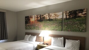 Habitación de hotel con dos camas, una mesita de noche con lámpara, y dos cuadros colgados en la pared de un campo y árboles.