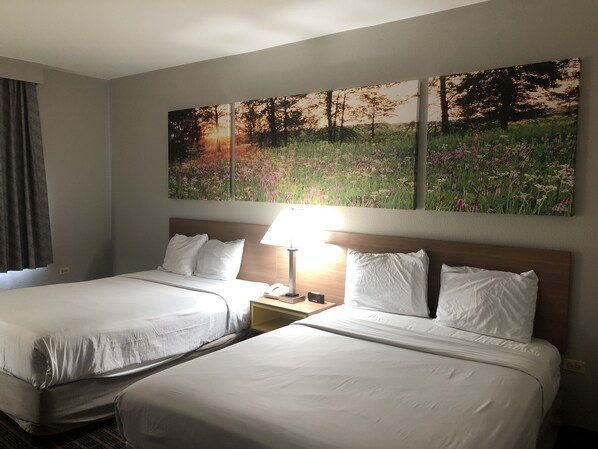 A hotel room with two beds, a nightstand with a lamp, and two wall-mounted paintings of a field and trees.