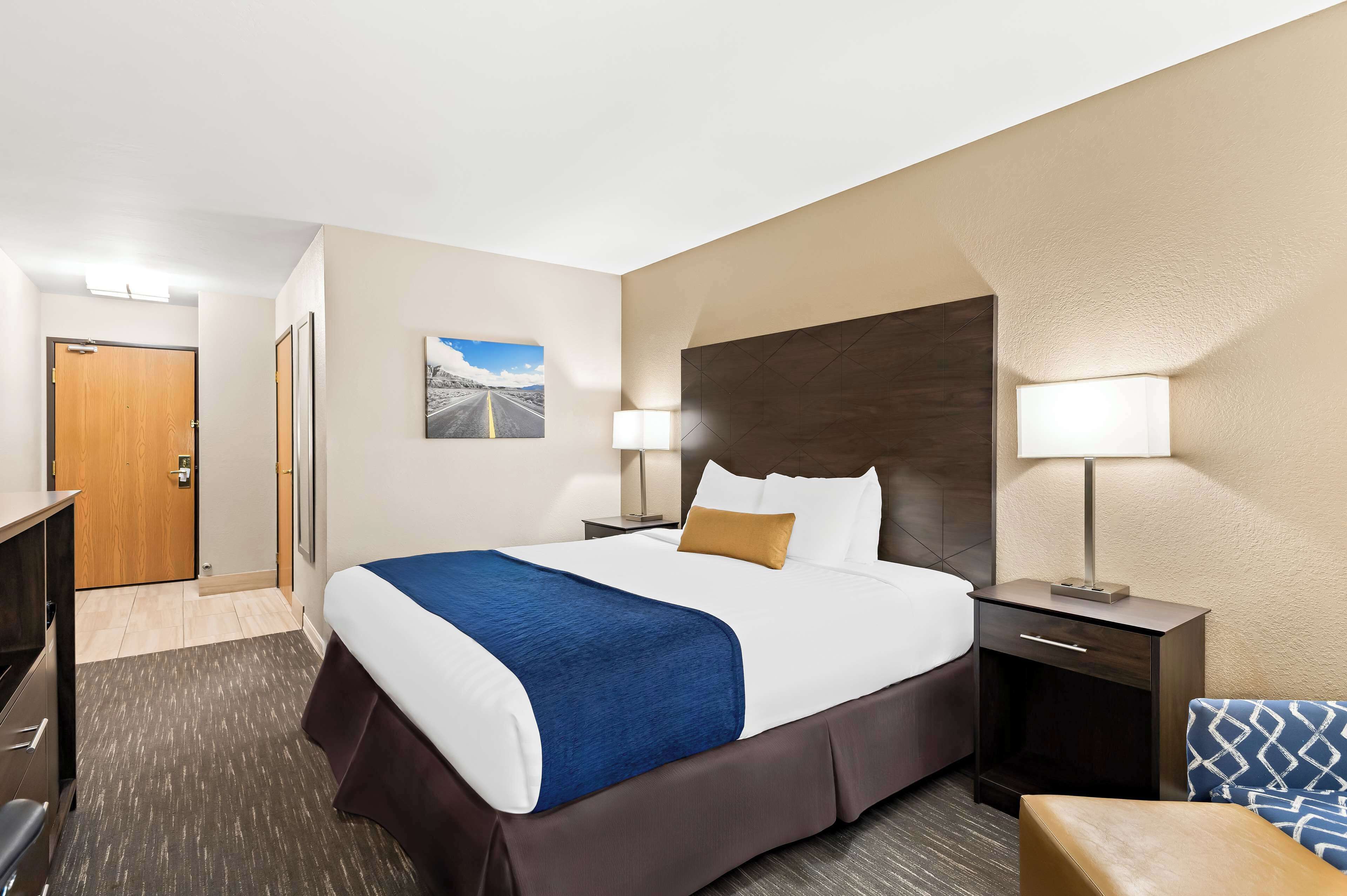A hotel room with a large bed, two bedside tables with lamps, a wooden door, and a framed picture on the wall.