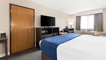 Habitación de hotel con una cama grande, televisor, escritorio con computadora, lámpara y ventana con cortinas.