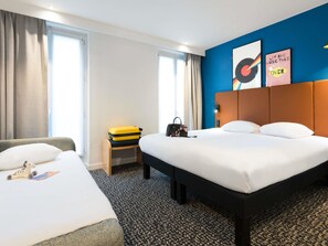 Ett hotellrum med en stor säng, en mindre säng, ett skrivbord med en resväska på och en vägg med två inramade tavlor.