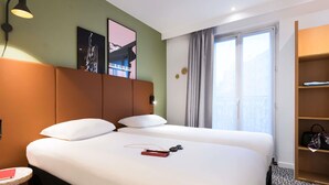 Habitación de hotel con dos camas, una mesita, una lámpara, un cuadro, una ventana con cortinas y un estante de madera con bolsos.