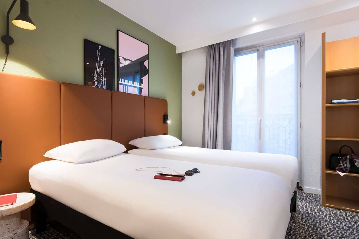 Hotelkamer met twee bedden, een tafeltje, een lamp, een schilderij, een raam met gordijnen en een houten plank met tassen.