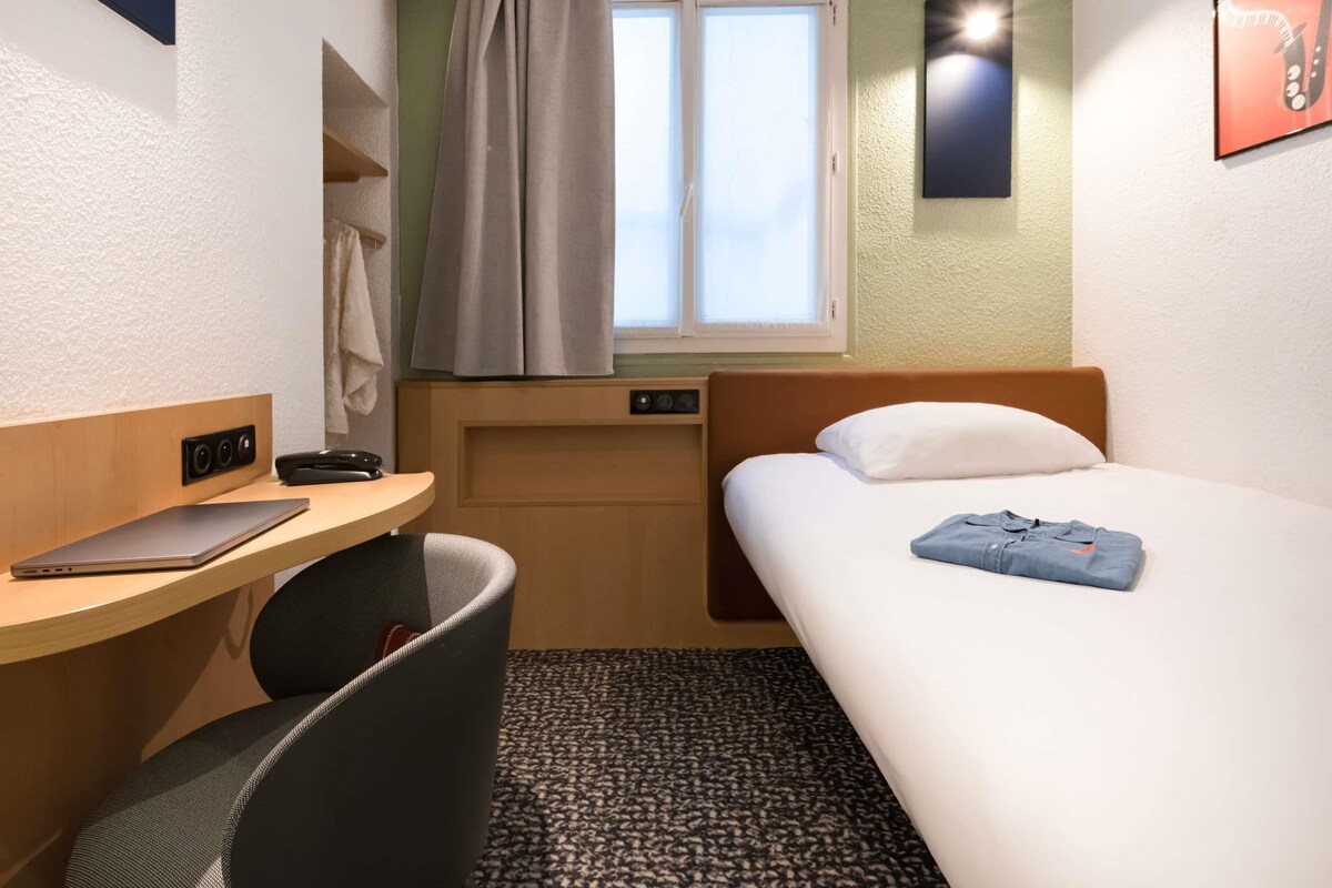 Een kleine hotelkamer met een eenpersoonsbed, een bureau met een laptop, een stoel, een raam met gordijnen en een wandlamp.