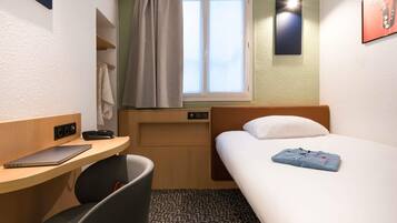 A compact hotel room with a single bed, a desk with a laptop, a chair, a window with curtains, and a wall-mounted light.