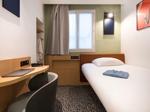 A compact hotel room with a single bed, a desk with a laptop, a chair, a window with curtains, and a wall-mounted light. - ibis Paris Gare Du Nord TGV (Paris)