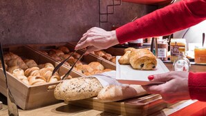 Un bufé con panes y pastelería variados, una persona sirviendo comida y una planta al fondo.