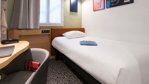A hotel room with a bed, a desk, a chair, a window with curtains, and wall art. - ibis Paris Gare Du Nord TGV (Paris)
