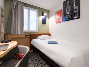 Et hotelværelse med seng, skrivebord, stol, et vindue med gardiner og billedkunst på væggen.