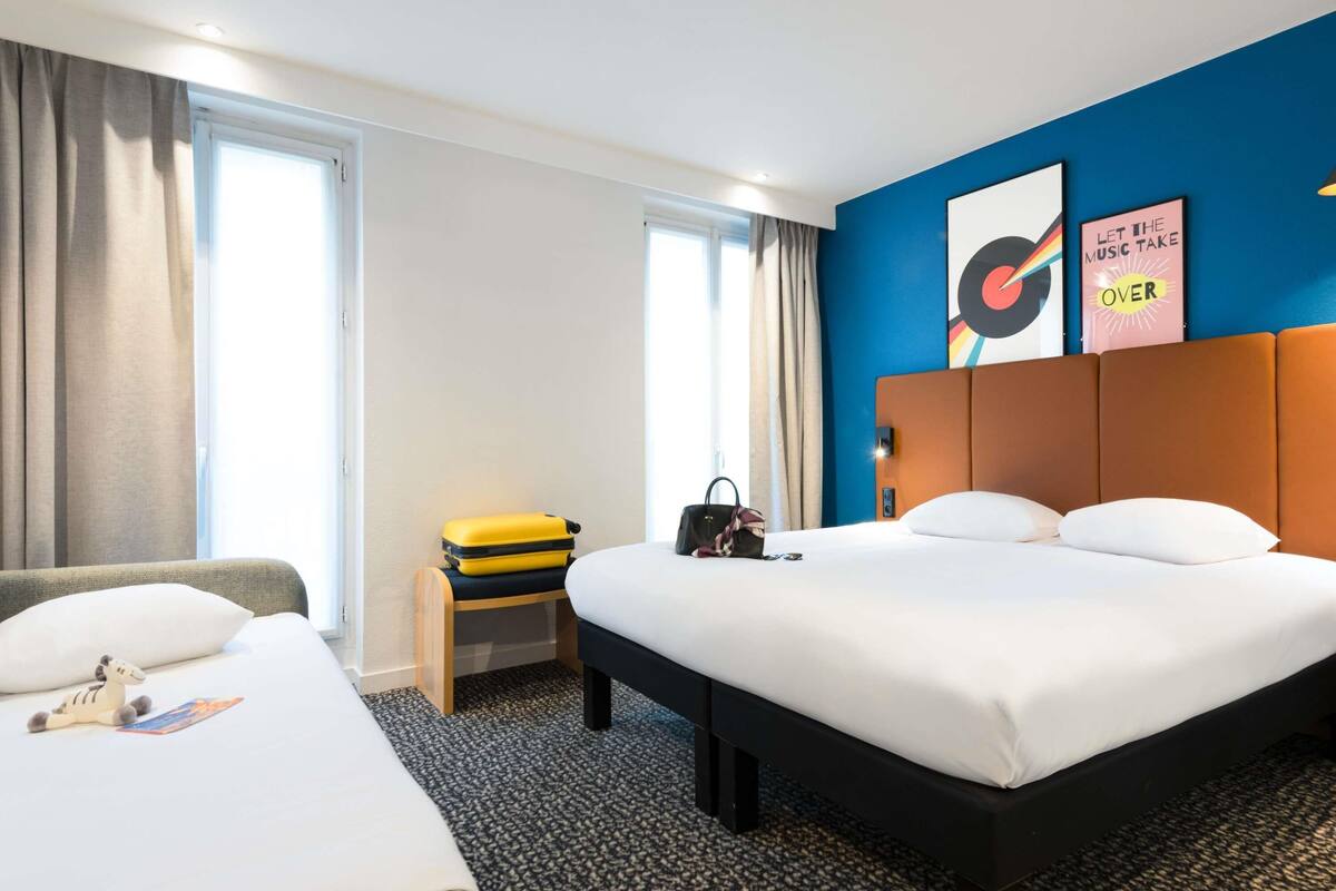 Een hotelkamer met een groot bed, een kleiner bed, een bureau met een koffer en een wand met twee ingelijste foto's.
