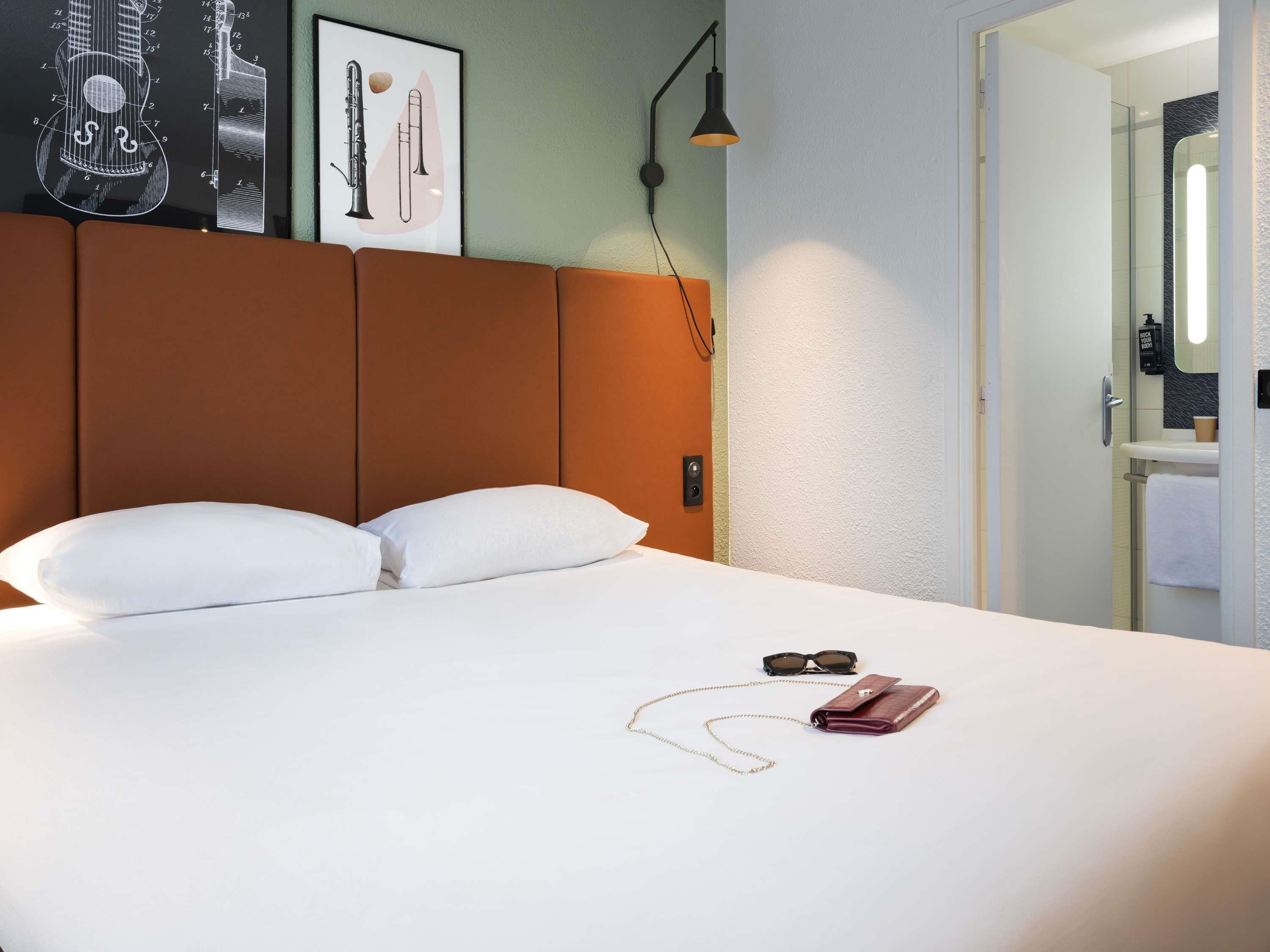 Une chambre d’hôtel avec un lit, des oreillers, un tableau représentant des instruments de musique, une table de chevet avec une lampe, et une salle de bain visible à travers une porte ouverte.
